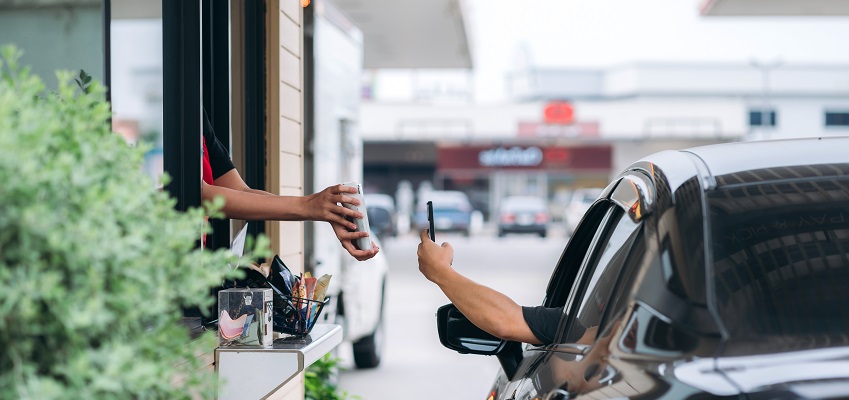 At a drive through window a customer completes transaction using his phone. No faces visible just hands. Perspective is from behind the customer car.