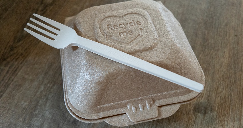 beige food container with plastic fork on top. sitting on wooden surface. container has text that reads 'recycle me'