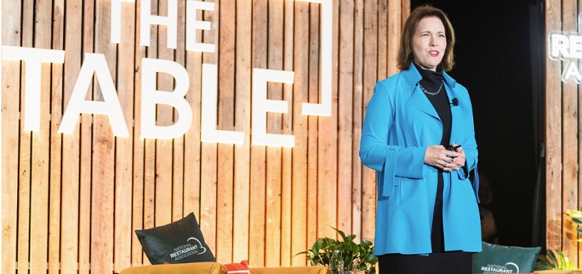 National Restaurant Association CEO Michelle Korsmo wearing blue blazer on stage in front of brown backdrop with Table logo